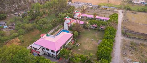 An aerial view of a residential area with multiple houses, a central building with a pink roof, and a dirt road.