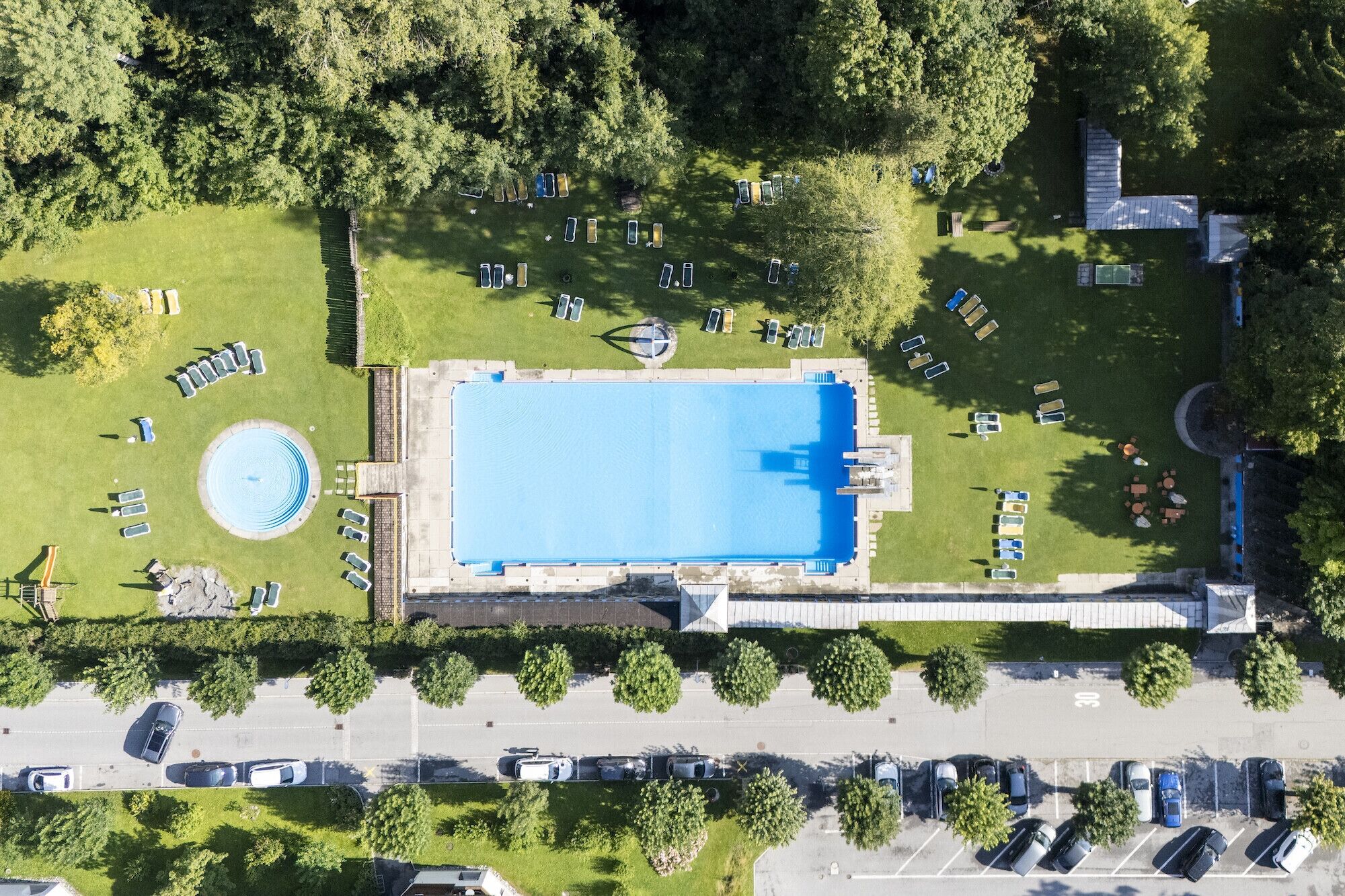 Eine Vogelperspektive auf ein Schwimmbecken, umgeben von Liegestühlen und einem Parkplatz.