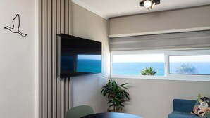 A modern living room with a round black table, two green chairs, a flat-screen TV mounted on the wall, a potted plant, a sofa with a colorful pillow, and a view of the ocean through a window with blinds. - Amazing Sea View Stylish 1BR | Safe Room on Floor (Herzliya)