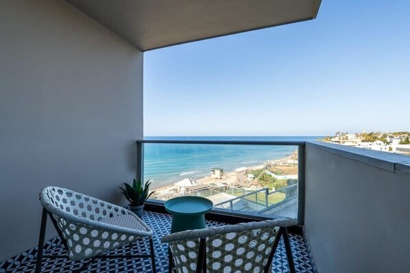 A balcony with two wicker chairs, a small table, and a view of the sea and buildings.