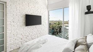 A modern bedroom with a white brick wall, a flat-screen TV mounted above the bed, a large window with a view of the outdoors, and a balcony with potted plants.