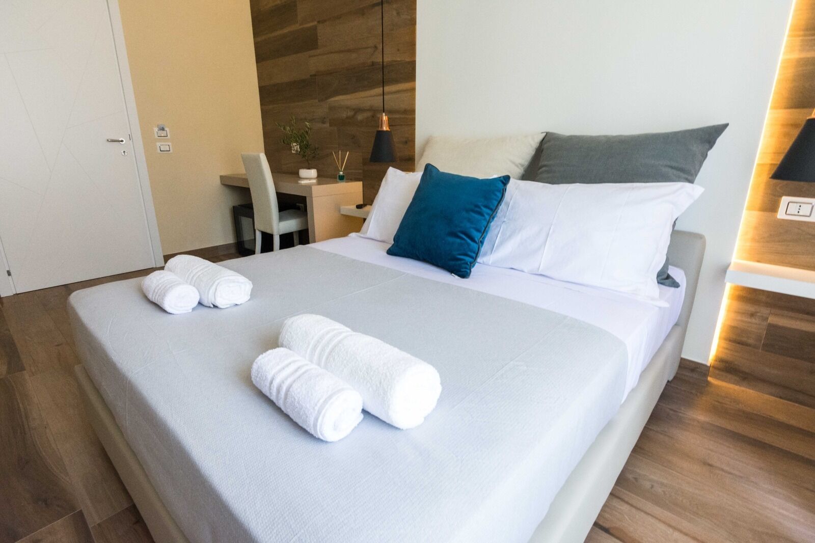 A neatly made bed with white linens and two rolled towels, a bedside table with a lamp, and a chair in a modern room with wooden flooring and a wooden accent wall.