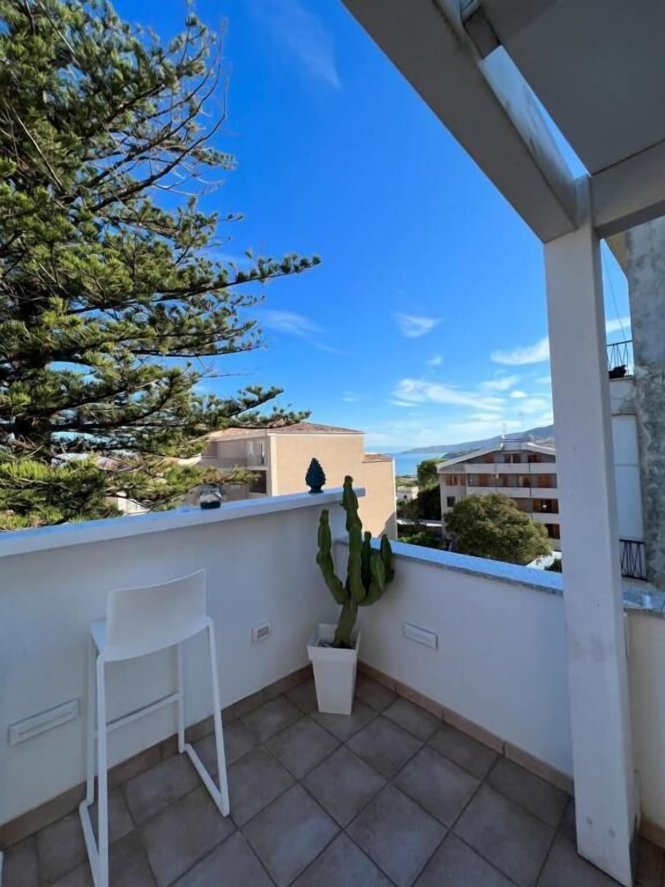 A balcony with a white chair, a potted cactus, and a view of buildings and trees.