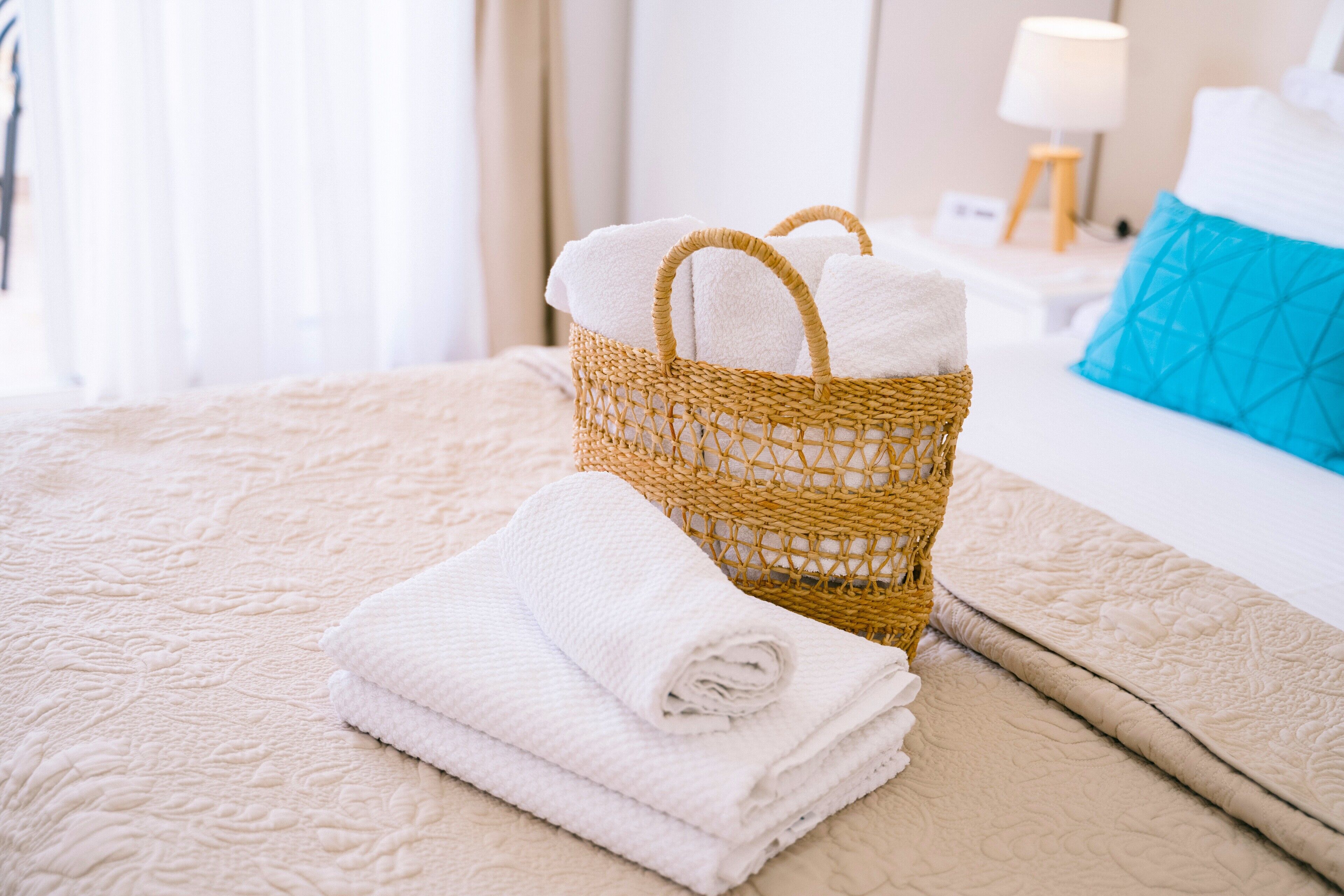 Un lit bien fait, recouvert d’une courtepointe beige, un panier en osier contenant des serviettes pliées et un oreiller bleu sur un lit blanc.