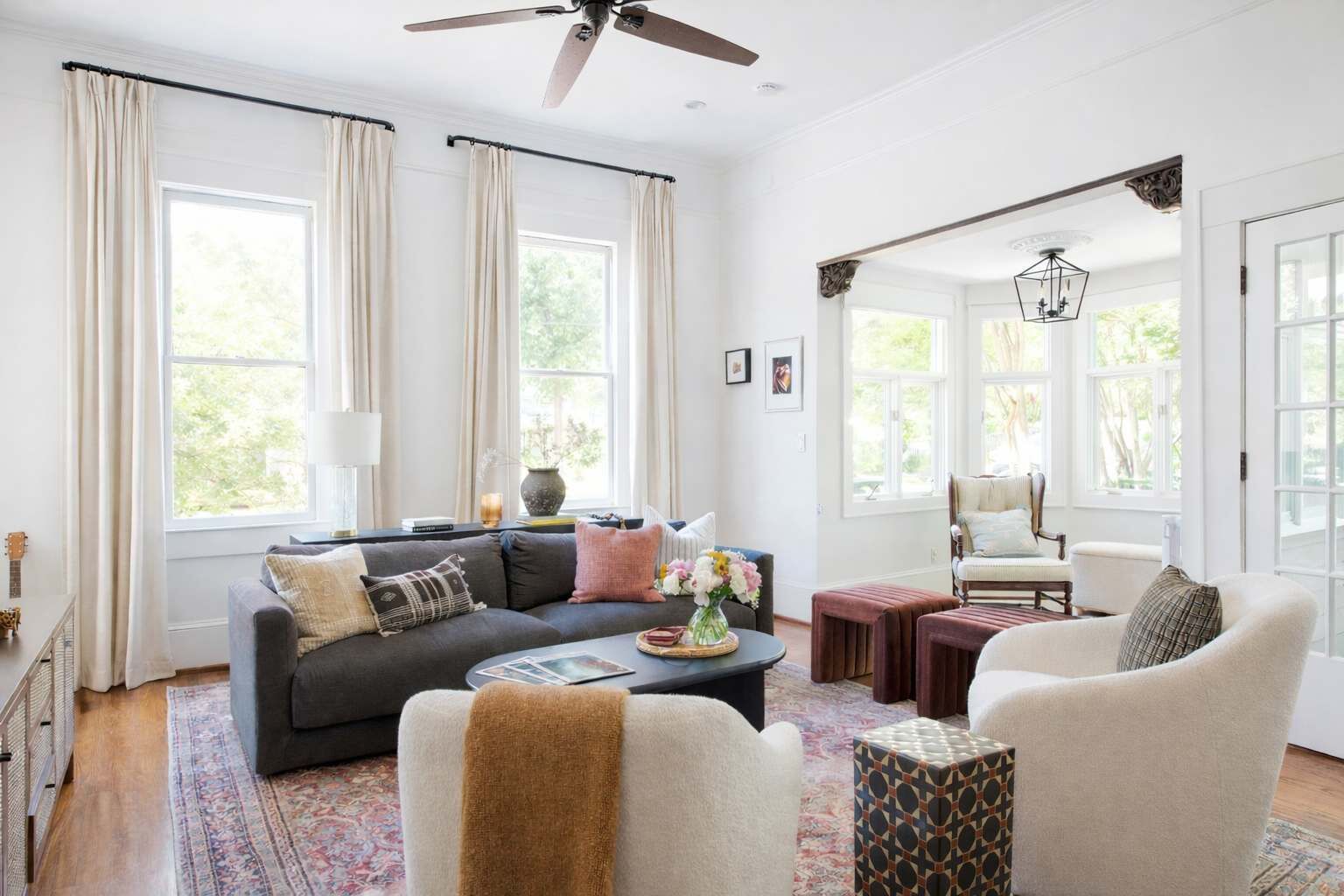 A spacious living room with a dark grey sofa, a white armchair, and a patterned ottoman. There is a coffee table with a vase of flowers and a book. The room has large windows with beige curtains, a ceiling fan, and a decorative light fixture.
