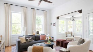 A spacious living room with a dark grey sofa, a white armchair, and a patterned ottoman. There is a coffee table with a vase of flowers and a book. The room has large windows with beige curtains, a ceiling fan, and a decorative light fixture. - Grant Grandeur (Atlanta)