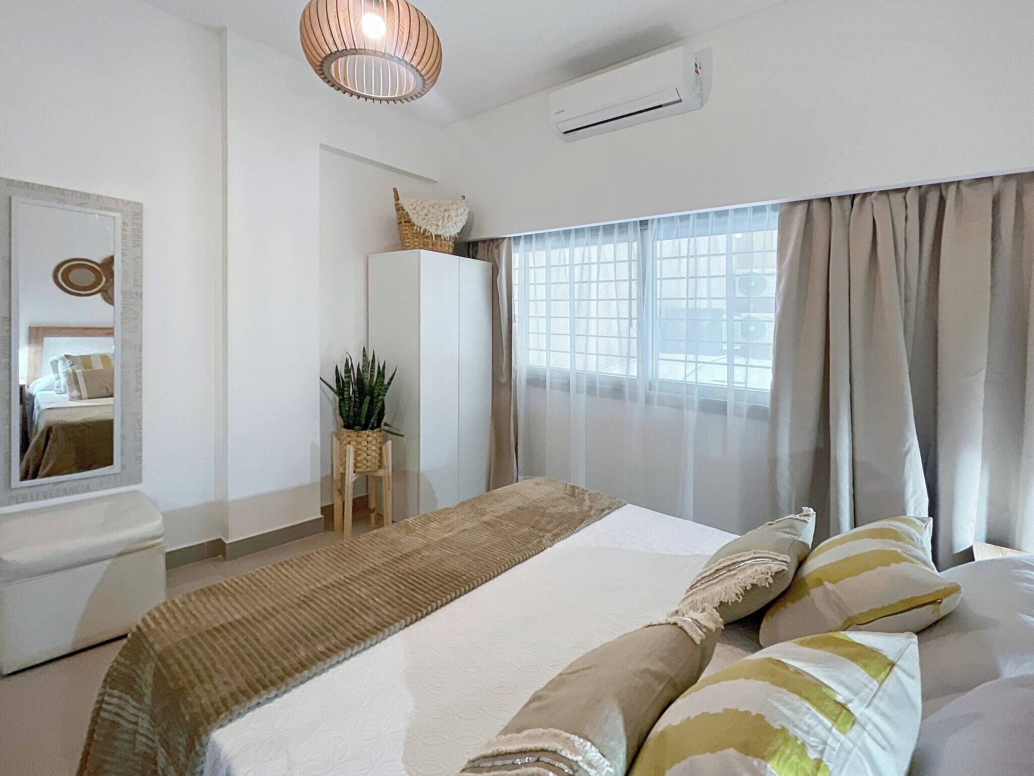 A modern bedroom with a large bed, a bedside table, a mirror, a potted plant, and a window with curtains.