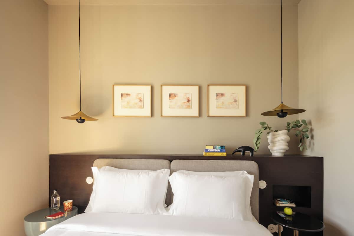 A hotel room with a bed, bedside tables, a nightstand, framed artwork, a vase with plants, and a bookshelf.