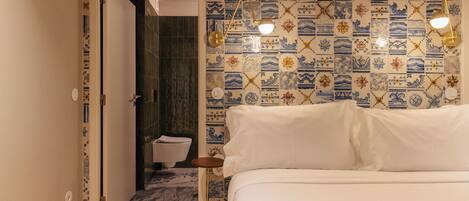 Quarto de hotel com uma cama, uma mesa de cabeceira, uma luminária fixada na parede e um banheiro visível através de uma porta aberta.