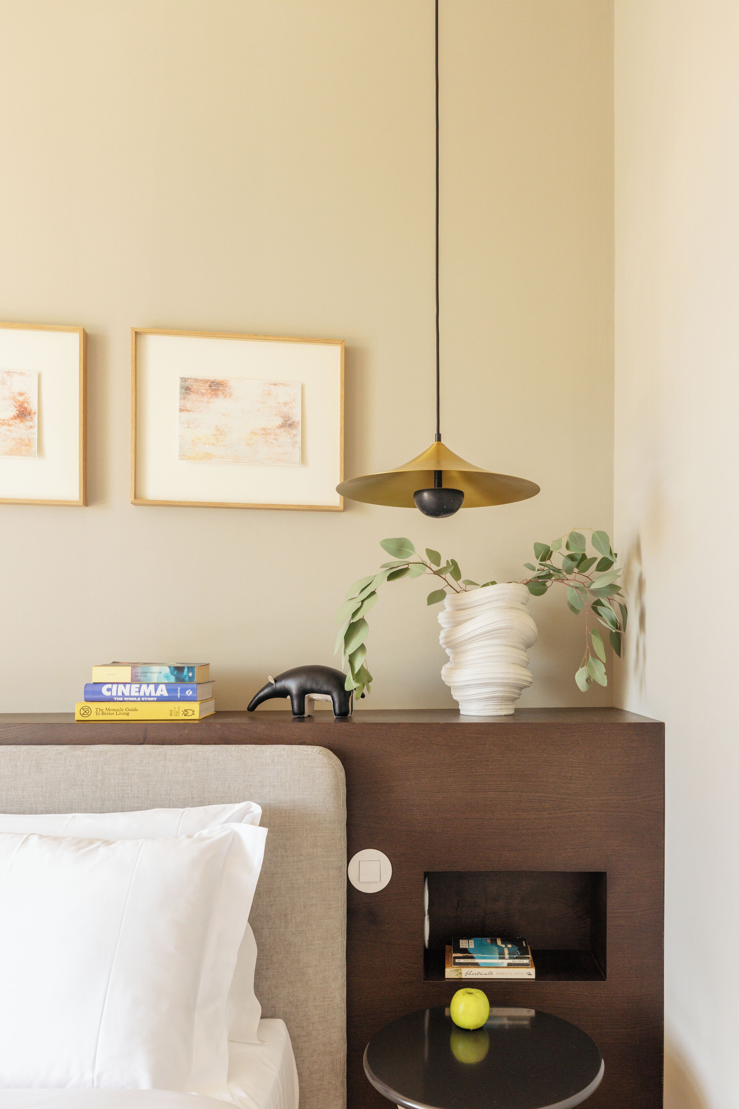 Een keurig opgemaakt hotelkamer met een bed, een nachtkastje met een lamp, ingekaderde foto's aan de muur en een klein plankje met boeken en decoratieve objecten.