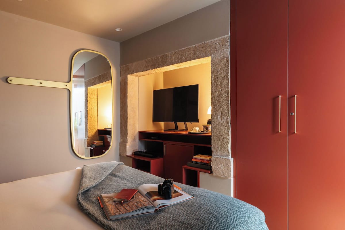A bedroom with a bed, a bedside table with a book and a camera, a mirror, a TV, and a wardrobe.