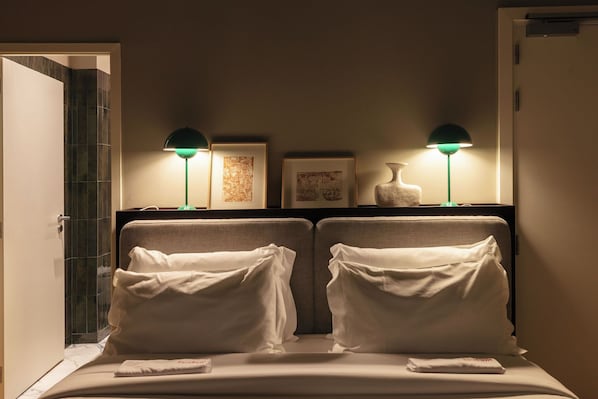 A hotel room with two beds, each with white linens and pillows, a nightstand with a lamp, and framed artwork on the wall.