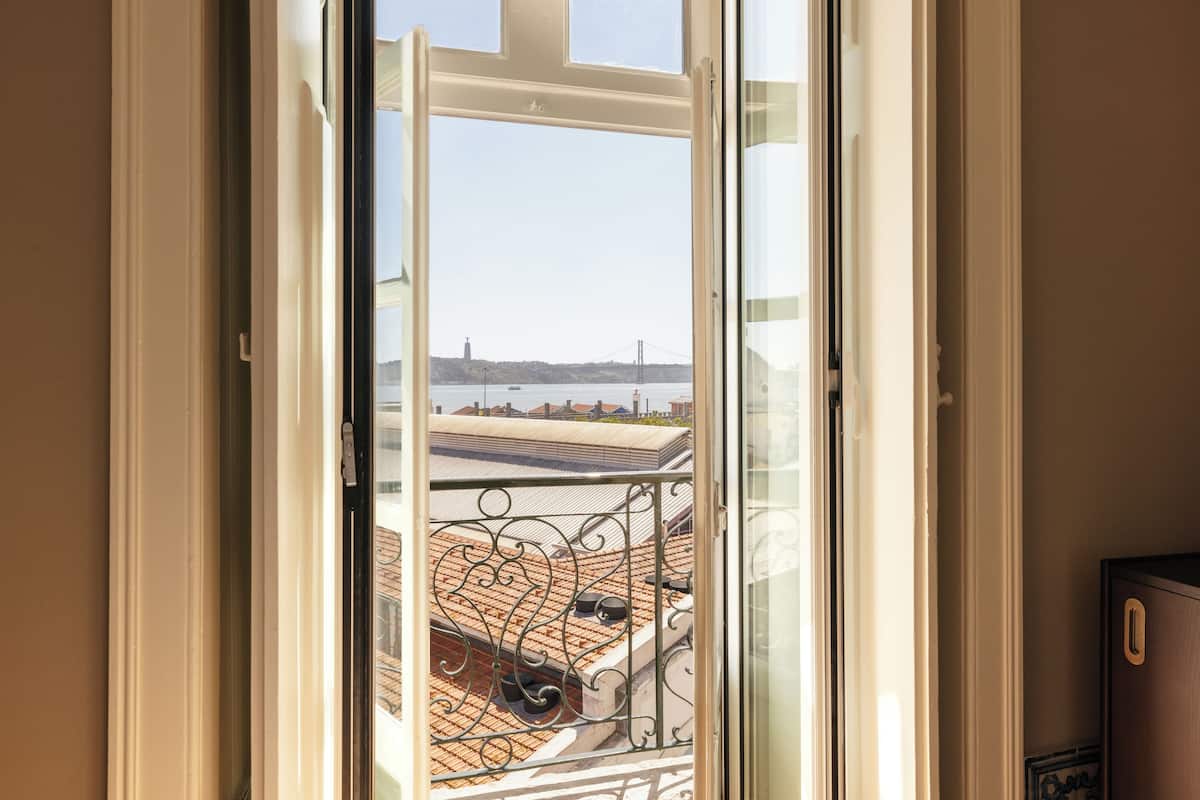 A view through a window with a metal railing, overlooking a building with a tiled roof and a distant bridge.