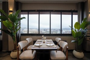 Uma área de refeições com uma mesa posta para quatro pessoas, plantas em vasos grandes e vista da cidade através de amplas janelas.