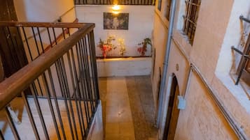 Una escalera estrecha con barandilla de madera y balaustrada de metal, que conduce a un rellano con un pequeño arreglo floral y un accesorio de luz montado en la pared.