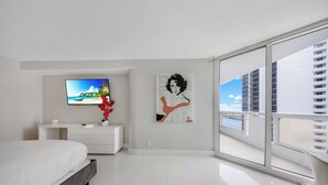 Une chambre moderne avec un lit, un téléviseur à écran plat, une commode blanche, un vase avec des fleurs rouges et une grande fenêtre donnant sur la ville.