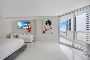 Une chambre moderne avec un lit, un téléviseur à écran plat, une commode blanche, un vase avec des fleurs rouges et une grande fenêtre donnant sur la ville.