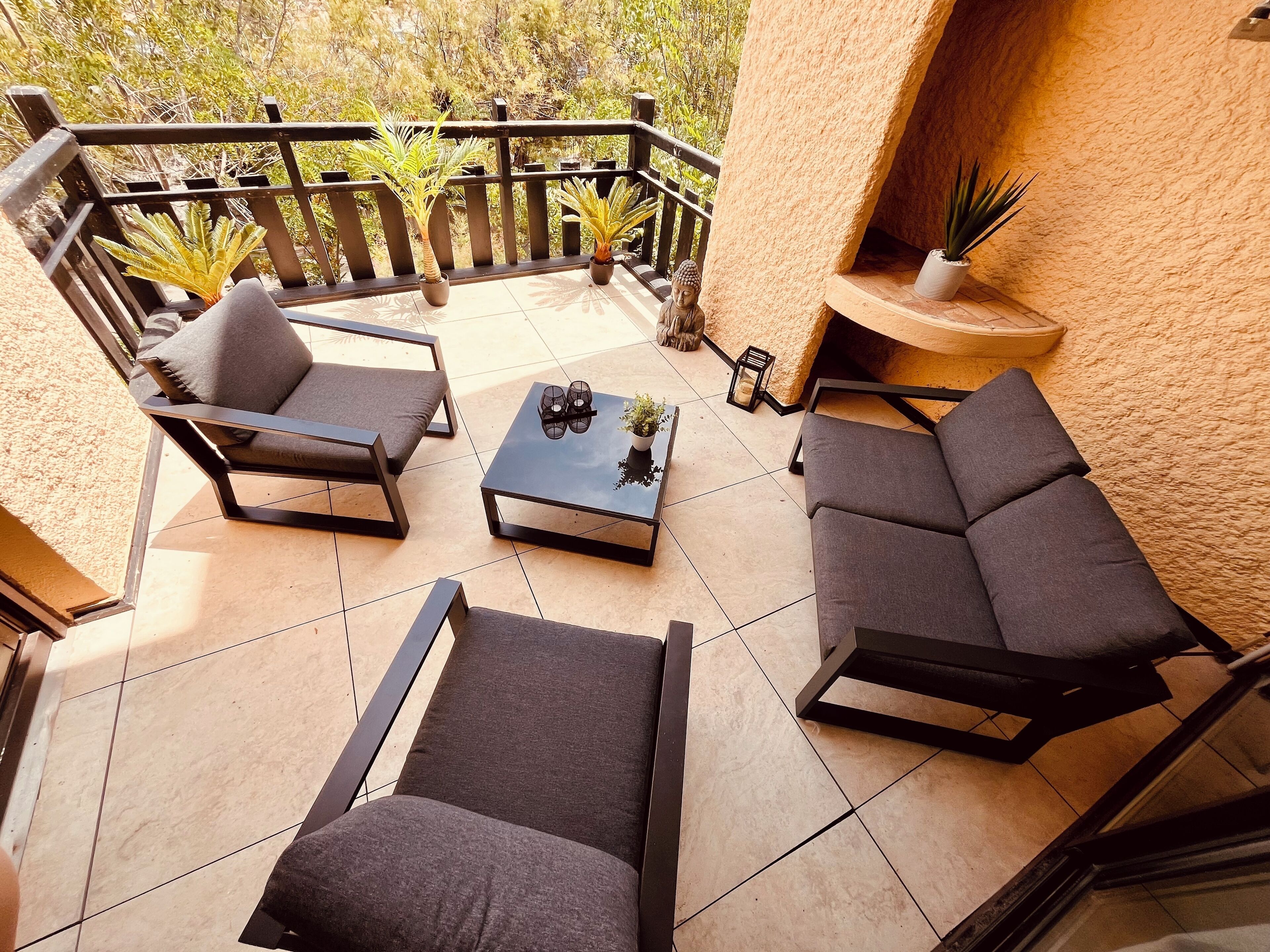 Ein sonniger Außenbereich mit Sitzgelegenheiten und modernen Möbeln, darunter zwei Sofas und ein Couchtisch, umgeben von Topfpflanzen und einer Buddha-Statue.