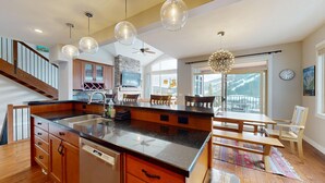 A spacious kitchen with a large island, wooden cabinets, and a Bosch dishwasher. There is a dining area with a table and chairs, and a view of mountains through large windows. - Luxurious, Village Center Home! (Sun Peaks)