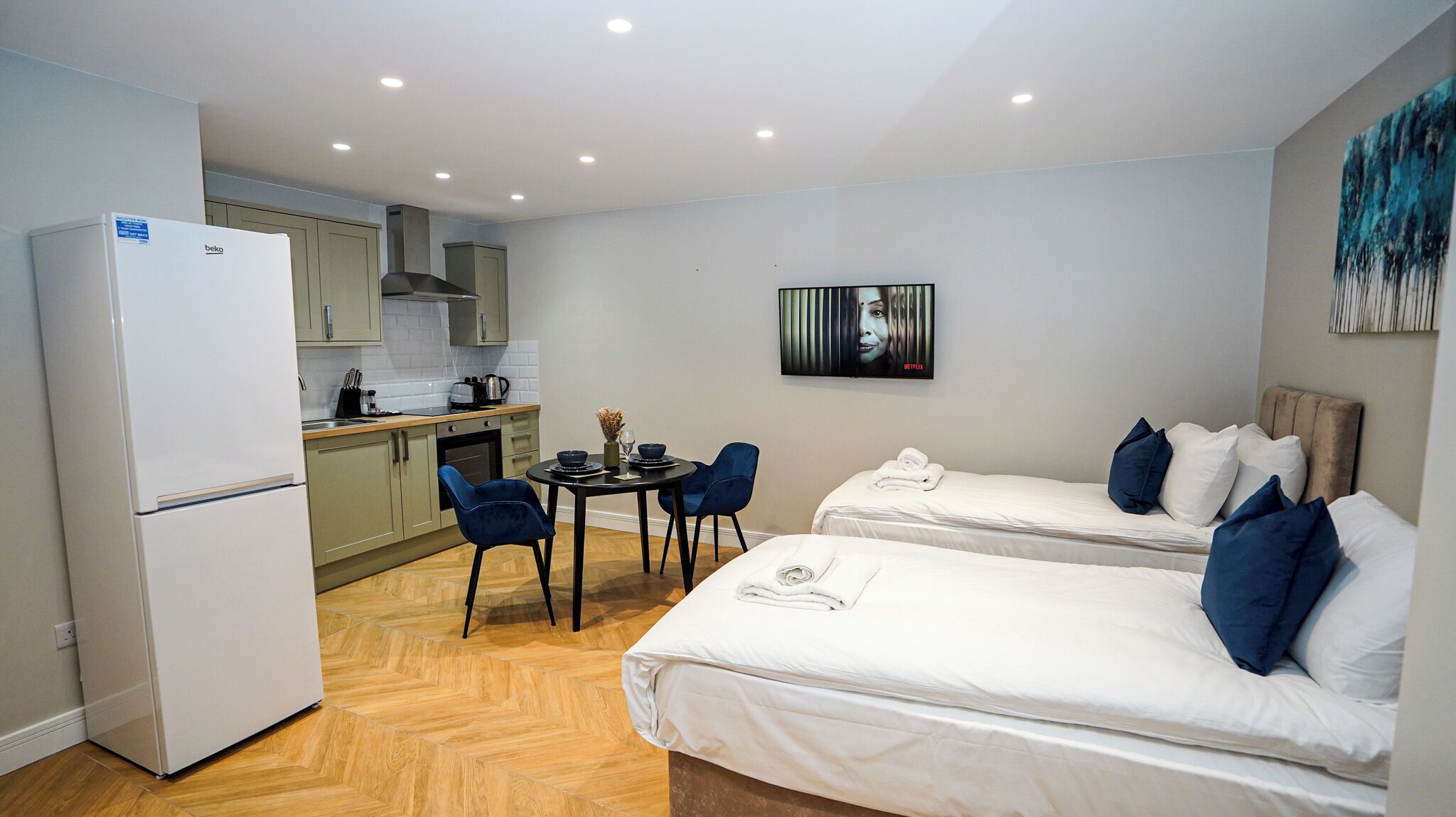 A compact hotel room with a kitchenette, two single beds, a dining table with chairs, and a painting on the wall.