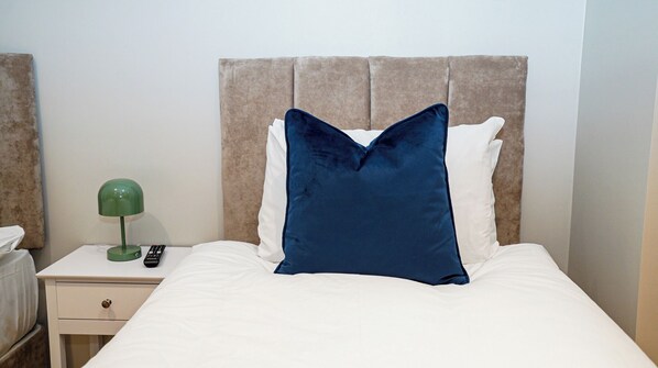 A neatly made bed with a white comforter and a large blue pillow, a green lamp on a bedside table, and a remote control. - Wisteria F2 - single beds (Slough)