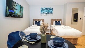 A hotel room with two beds, a dining table with blue tableware, a vase with dried flowers, and a TV displaying 'Sonic 2'. - Wisteria F2 - single beds (Slough)