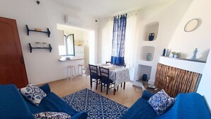 A living room with blue sofas and patterned cushions, a dining area with a table and chairs, and a kitchen area with shelves and a window. - Rosa's House Historic Center (Orosei)