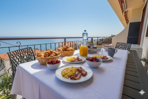 Een ontbijttafel met croissantjes, aardbeien en roerei op een balkon met uitzicht op zee.