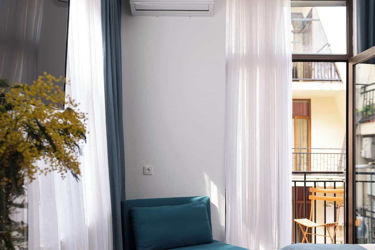 A modern hotel room with a wooden desk, a teal sofa, a bed with a blue bedspread, and a balcony with a view of the city.