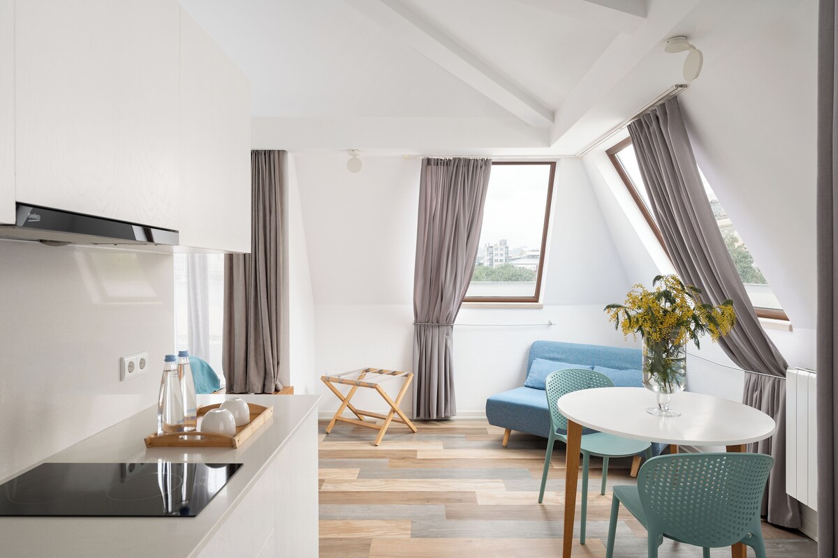 A modern kitchen with a built-in stove, a dining area with a round table and chairs, and a living space with a sofa and a view of the city.