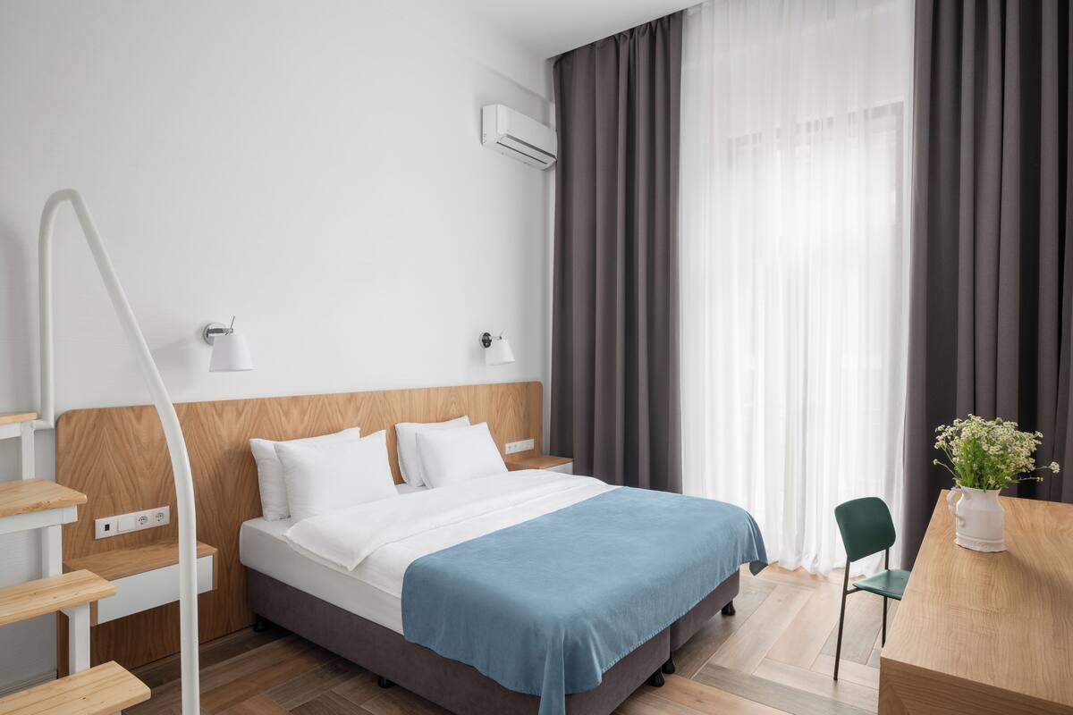 A modern hotel room with a bed, bedside tables, a desk, a chair, and a window with curtains.