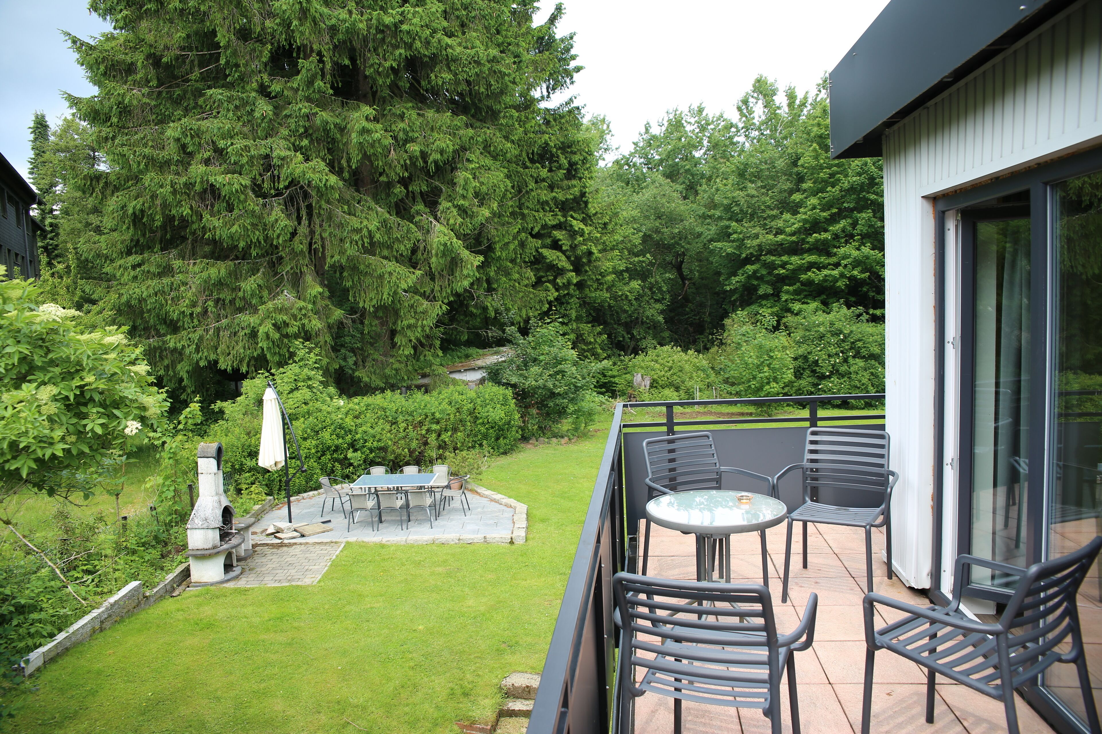 Ein Balkon mit Tisch und Stühlen, ein Kamin aus Stein und Blick auf einen üppigen Garten sowie hohe Bäume.