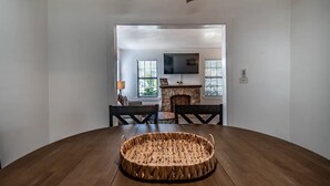 A dining area with a round wooden table and chairs, a woven basket in the center, and a stone fireplace in the background. - Blue Wave Cottage Sum (Corpus Christi)