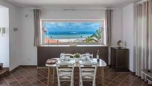 A dining area with a table set for two, a window with a view of the beach, and a small cabinet with a decorative item. - 3997 Villa Thalissa by Barbarhouse (Taranto)