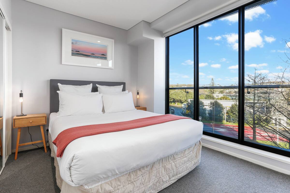 A modern bedroom with a large bed, a nightstand, a bedside lamp, a framed picture on the wall, and a large window with a view of the city.