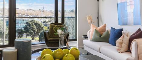 A modern living room with a large window, a sofa with colorful cushions, a bowl of apples, and a vase with flowers.