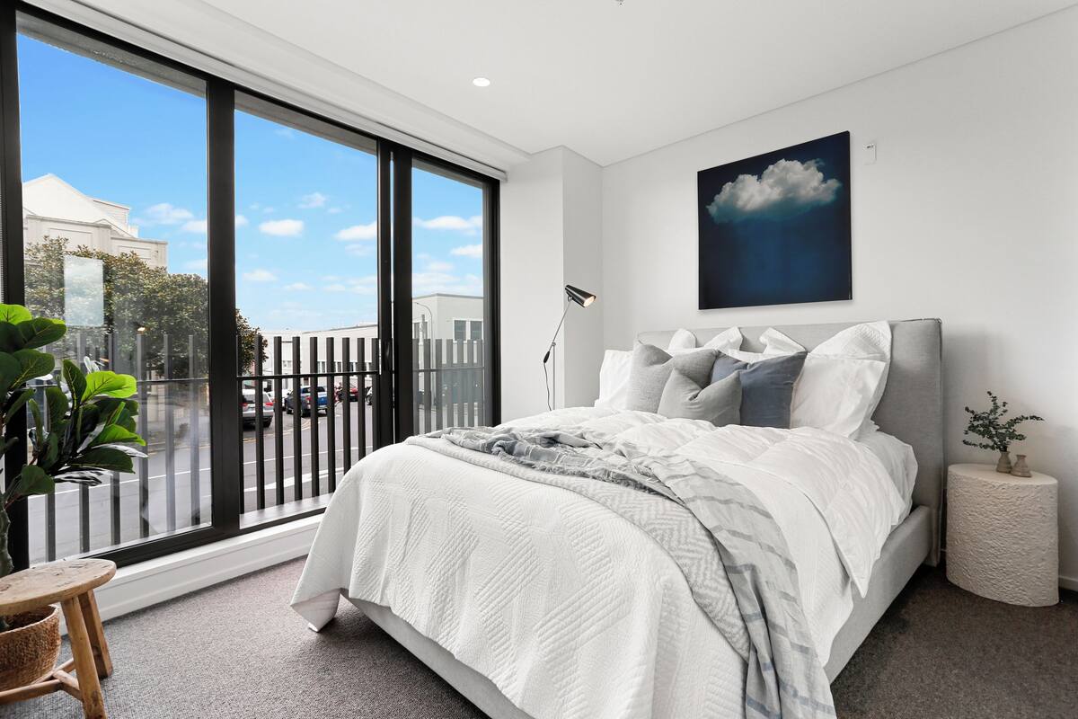 A modern bedroom with a large window, a bed with white bedding, a grey headboard, and a wall-mounted painting of a cloud.