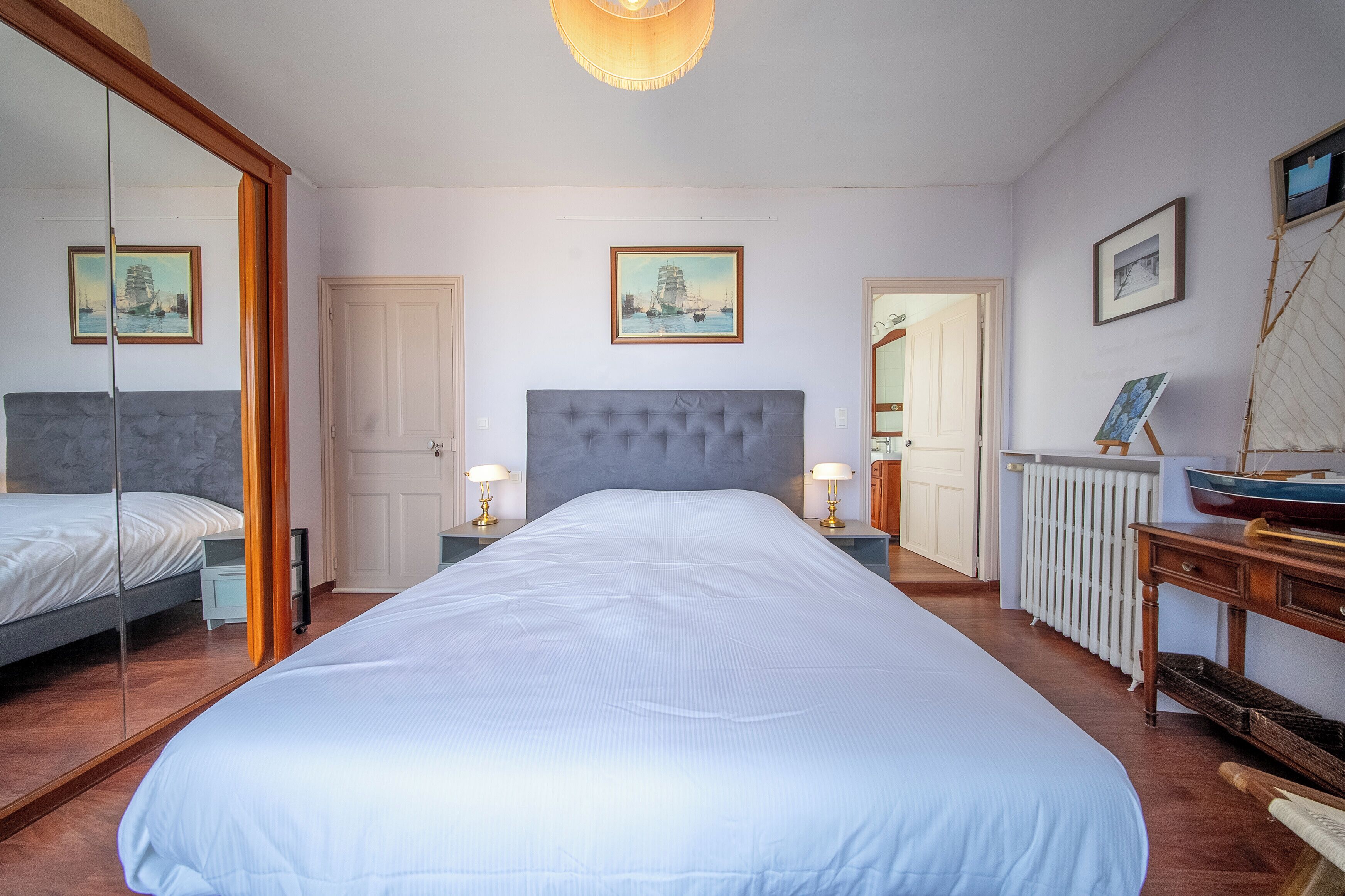Un dormitorio con una cama grande, dos mesitas de noche con lámparas, un espejo, un aparador de madera y un radiador.