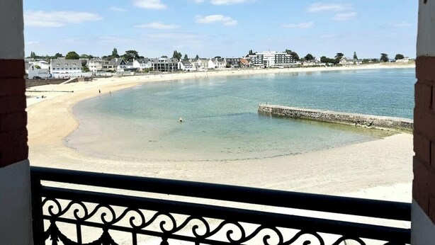 Vista a la playa desde un balcón con barandilla, cielo despejado y edificios al fondo.