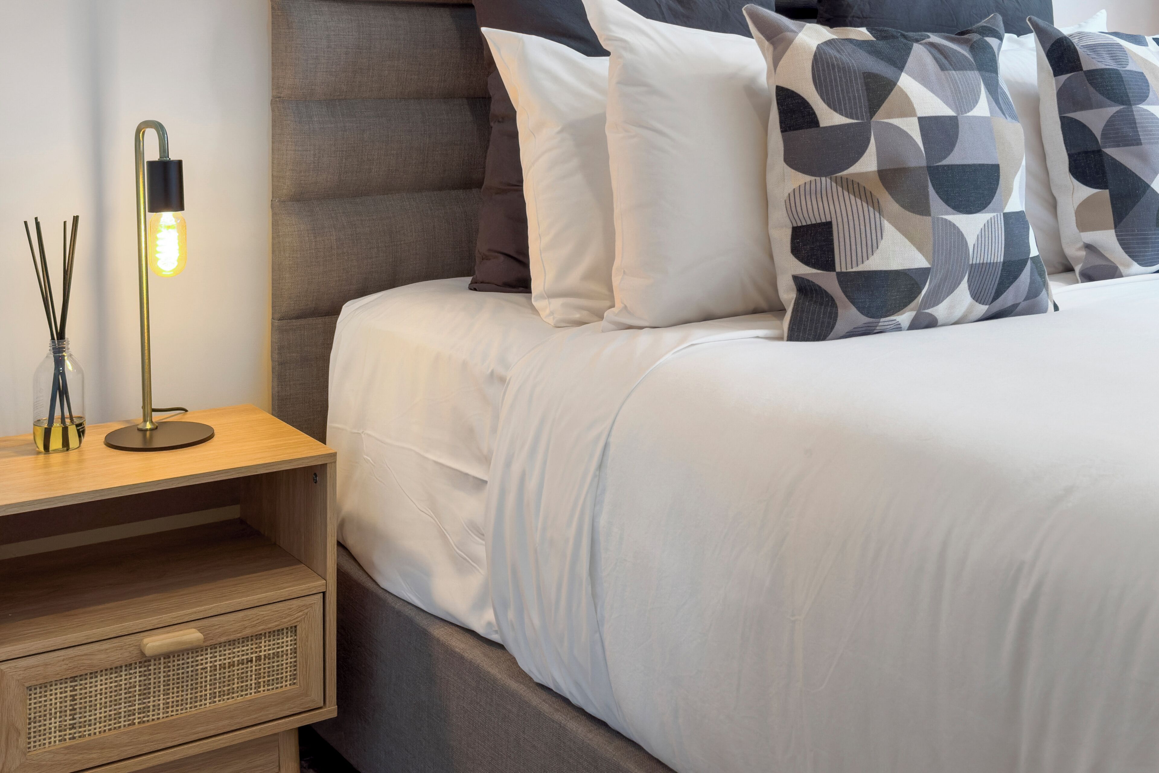 A neatly made bed with white bedding and geometric patterned pillows, a bedside table with a lamp, and a drawer unit.