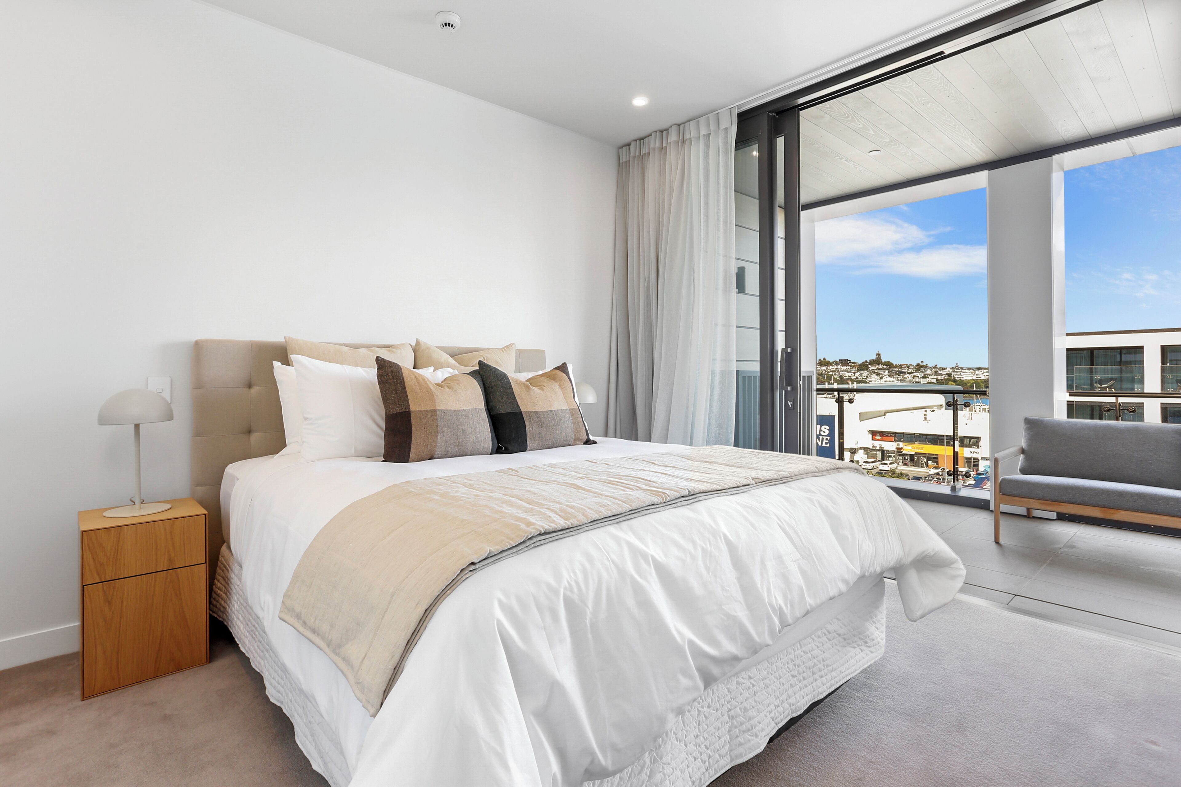 A modern bedroom with a large bed, a nightstand, a sofa, and a view of the city through large windows.