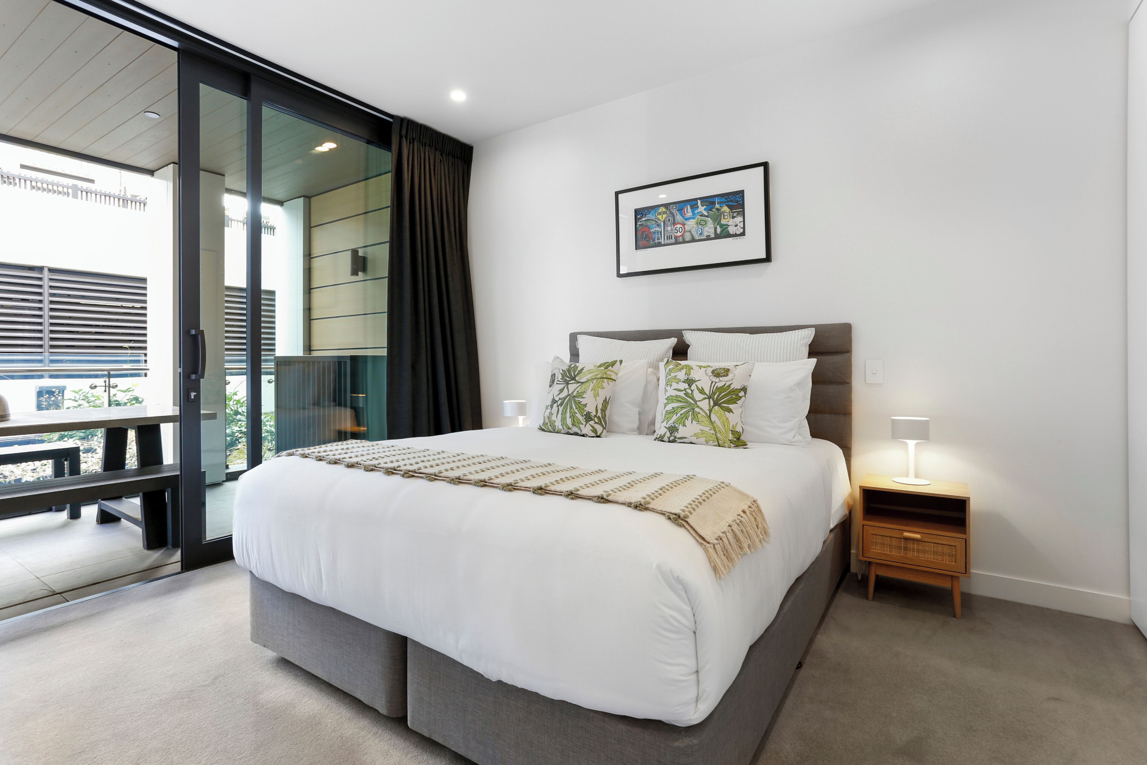 A bedroom with a large bed, a bedside table with a lamp, a framed picture on the wall, and a sliding glass door leading to an outdoor area.