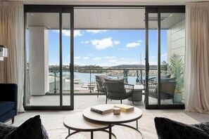 A modern living room with a large sliding glass door leading to a balcony with a view of a marina and hills. - Wynyard Quarter Apartments by Urban Rest (Auckland)