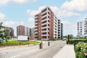 Un edificio residencial de varios pisos con balcones, un sendero pavimentado y un área ajardinada con vegetación.