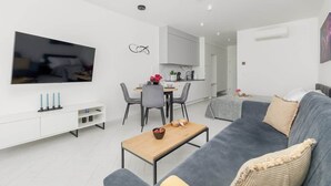 Un salon moderne avec un canapé gris, une table basse en bois et un téléviseur à écran plat fixé au mur. On y trouve également un coin repas avec une table et des chaises, ainsi qu’une cuisine équipée de placards et d’un plan de travail.