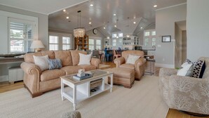 A spacious living room with a high ceiling, a central chandelier, and a kitchen area visible in the background. - Stunning Views With Pool, Family Room, Resort Perks (Corolla)