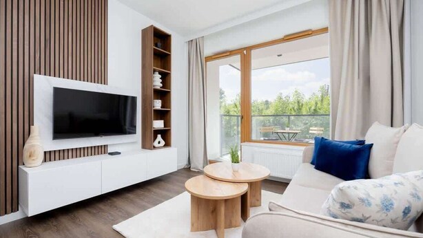 A modern living room with a flat-screen TV, wooden sideboard, two round wooden tables, a sofa with floral cushions, and a balcony with a view of greenery. - Modern Apartment | Parking | Close to Metro (Warszawa)