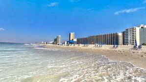 A beach with clear waters, a sandy shore, and a row of high-rise buildings in the background. - Gulf Front at Phoenix VII 7916 | Huge Indoor/Outdoor Pools | Amenities Galore (Orange Beach)