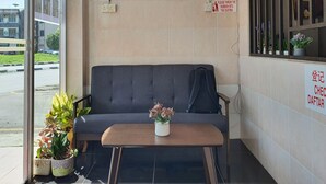 Un área de espera con un sofá, una mesita con un florero, plantas en macetas y una ventana con vista a la calle.