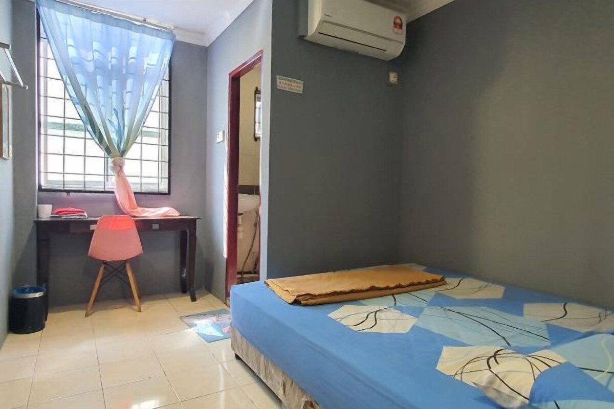 Una habitación pequeña con una cama, un escritorio y una silla. Tiene una ventana con cortina, un equipo de aire acondicionado y una puerta.